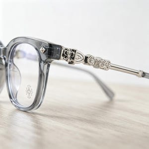 May include: Gray eyeglasses with a decorative silver metal arm. The frames are a translucent gray, and the arms feature ornate cross and floral designs. The lenses are clear, and the overall style is modern and stylish.