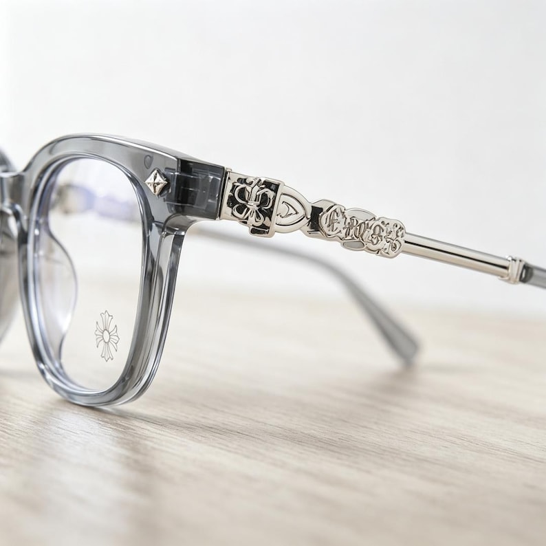 May include: Gray eyeglasses with a decorative silver metal arm. The frames are a translucent gray, and the arms feature ornate cross and floral designs. The lenses are clear, and the overall style is modern and stylish.