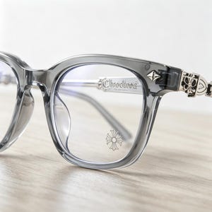 May include: Gray square-framed eyeglasses with silver accents. The temples feature ornate silver detailing and the brand name "Chroclivos" is etched on the frame. A decorative emblem is visible on the lower right lens.