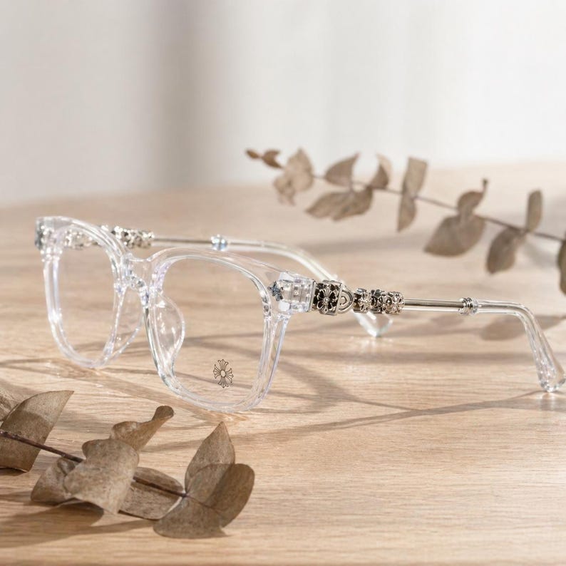 May include: Clear eyeglasses with square frames and decorative embellishments on the temples. The temples are silver with black and clear accents. The lenses are clear, and the glasses are resting on a wooden surface.