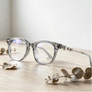 May include: Clear gray eyeglasses with a square frame and silver accents. The temples feature decorative silver embellishments. The lenses are clear, and the glasses are set on a wooden surface.