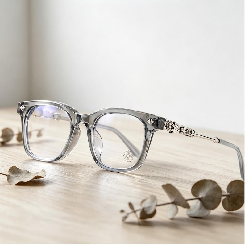 May include: Clear gray eyeglasses with a square frame and silver accents. The temples feature decorative silver embellishments. The lenses are clear, and the glasses are set on a wooden surface.