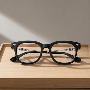 May include: Black eyeglasses with a square frame and silver accents. The temples feature ornate silver detailing, and the lenses are clear. The glasses have a modern design and are set on a wooden tray.