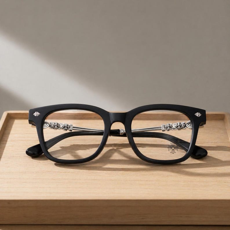 May include: Black eyeglasses with a square frame and silver accents. The temples feature ornate silver detailing, and the lenses are clear. The glasses have a modern design and are set on a wooden tray.
