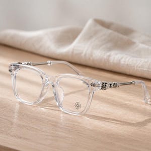 May include: Transparent square-framed spectacles with silver detailing on the arms. The lenses are clear, and a small circular emblem is visible on the lower right lens. The frames are clear.