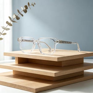 Puede incluir: Gafas transparentes de montura cuadrada con detalles plateados decorativos en las patillas y un pequeño emblema en la lente inferior derecha. Las gafas se exhiben sobre una pila de estantes de madera sobre un fondo azul claro.