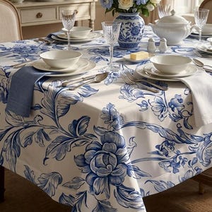 Pode incluir: Uma toalha de mesa branca com um padrão floral azul sobre uma mesa de jantar. A mesa está posta com tigelas e pratos de cerâmica branca, copos de cristal transparente, talheres de prata, um guardanapo azul, um branco e um vaso azul e branco.