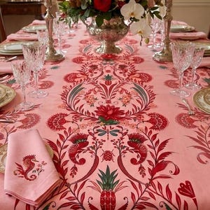 May include: A pink tablecloth featuring a repeating pattern of red and green botanical motifs, including pineapples, flowers, and insects. A matching pink napkin with a red seahorse embroidery is placed on the table, which is set with crystal glasses.