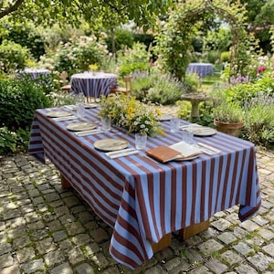 May include: A rectangular tablecloth featuring vertical stripes in alternating shades of light blue and dark brown. The fabric covers a wooden table set with clear glass tumblers, white plates, silverware, and a central floral arrangement.