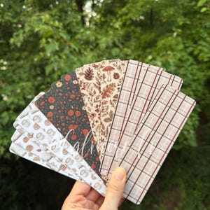 May include: A set of rectangular bookmarks fanned out, featuring various patterns including a white background with coffee cups, a dark green floral print, a cream background with botanical illustrations of mushrooms and pinecones, and a beige plaid design.