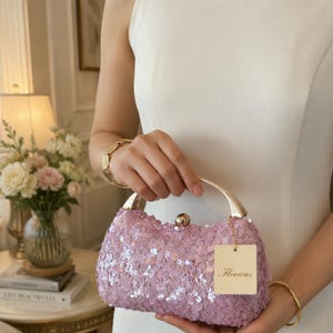Op de afbeelding: Een lichtpaarse clutch bedekt met glinsterende pailletten, voorzien van een halfrond handvat van goudkleurig metaal en een centrale sluiting. Een rechthoekig beige label met de merknaam Flovious is aan het handvat bevestigd.
