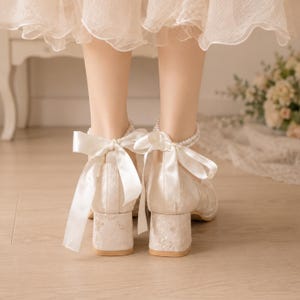 May include: Off-white block heel shoes featuring a textured floral fabric finish, a delicate pearl ankle strap, and a large satin ribbon bow tied at the back of the heel. The shoes have a sturdy, low block heel and a closed-back design.