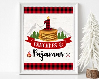 Könnte beinhalten: Gerahmter Druck mit rustikalem Thema. Das Design zeigt einen Stapel Pfannkuchen mit einer Kerze mit der Nummer eins, Berge und die Worte "Pancakes & Pajamas". Rote und schwarze Büffel-Plaid-Ränder umranden die Ober- und Unterseite.