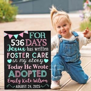 May include: A black chalkboard sign with white text that reads "FOR 536 DAYS Jesus has written FOSTER CARE in my story Today He Wrote ADOPTED Emily Kate Wilson AUGUST 24, 2025". The sign is decorated with pink and blue flags. A young girl in blue overalls is standing in front of the sign.