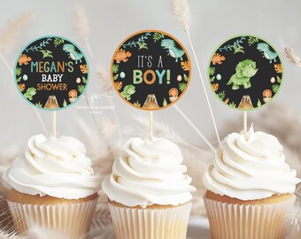 Könnte beinhalten: Drei Cupcakes mit weißem Zuckerguss und dekorativen Tortenauflegern. Die runden Aufleger mit schwarzem Hintergrund zeigen Dinosaurier-Illustrationen und die Texte "Megan's Baby Shower" und "It's a Boy!"