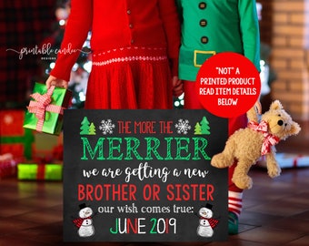 May include: A chalkboard sign with the text "The More The Merrier" in green letters. The sign also says "We are getting a new brother or sister, our wish comes true: June 2019". The sign is decorated with snowflakes and Christmas trees. Two children are holding gifts in front of the sign.