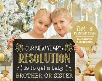May include: A black chalkboard sign with gold lettering announcing a new baby. The sign reads "Our New Year's Resolution is to get a baby Brother or Sister. Mommy is due: June 2024". Two children are holding the sign in front of a Christmas tree.