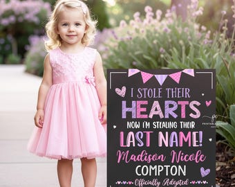 May include: A young girl in a pink dress and purple sneakers stands next to a chalkboard sign. The sign reads "I STOLE THEIR HEARTS NOW I'M STEALING THEIR LAST NAME!" with a name and adoption date. The sign is decorated with hearts and pennants.