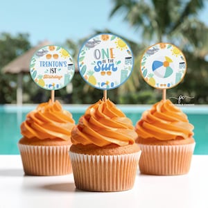 Może przedstawiać: Trzy babeczki z pomarańczowym lukrem i dekoracyjnymi topperami. Toppery są okrągłe z niebiesko-pomarańczową obwódką, z letnimi ilustracjami i tekstem "Trenton's 1st Birthday" i "One in the Sun".