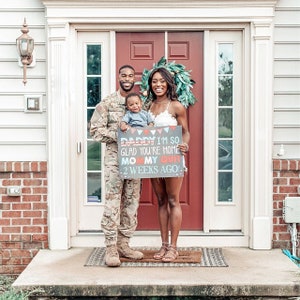 Puede incluir: Una familia de tres personas est&aacute; de pie frente a su casa, sosteniendo un cartel que dice "Pap&aacute;, estoy tan contento de que est&eacute;s en casa, Mam&aacute; renunci&oacute; hace 2 semanas!" El padre lleva un uniforme militar, la madre lleva un vestido blanco y el ni&ntilde;o lleva una camisa azul.