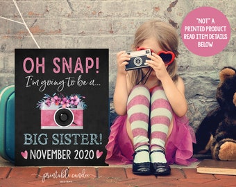 May include: A black chalkboard sign with pink and white text that reads "OH SNAP! I'm going to be a... BIG SISTER! NOVEMBER 2020". The sign features a pink and white camera with pink flowers on top. A young girl is holding a camera in front of her face and wearing pink heart-shaped sunglasses.