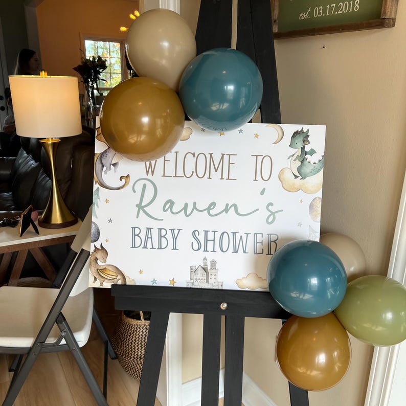 May include: A baby shower sign with a dragon design and the text "Welcome to Raven's Baby Shower". The sign is decorated with gold, teal, and brown balloons.