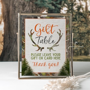 May include: A sign with a camouflage background and antlers, reading "Gift Table" in black text with orange outlines. The sign also says "Please leave your gift or card here" and "Thank you!" in black text.