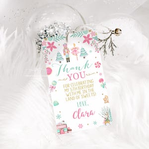 May include: A rectangular gift tag with a festive design. The tag features the words "Thank You" and "Clara" in a script font, along with illustrations of a nutcracker, ballerina, and sweets. The tag is decorated with pink and teal accents.