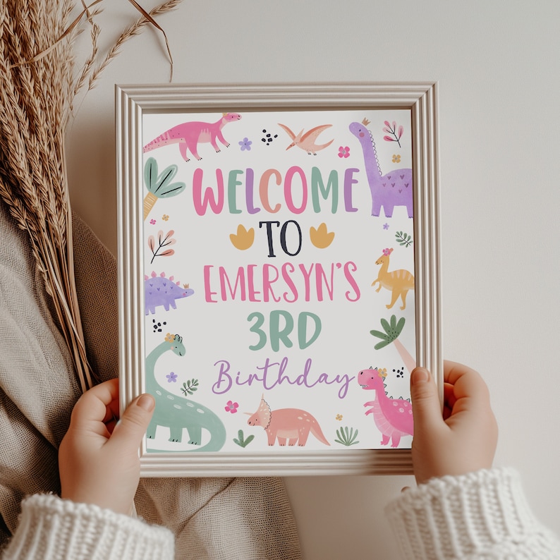 Puede incluir: Un cartel enmarcado en blanco con un tema de dinosaurio rosa, verde y morado. El cartel dice "Bienvenido a Emersyn's 3rd Birthday".