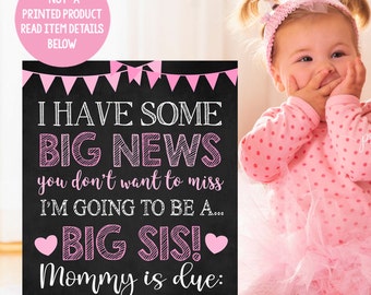 Op de afbeelding: Een zwart krijtbord met roze en witte letters kondigt een baby aan. Het bord staat er: "Ik heb groot nieuws dat je niet wilt missen Ik ga een... Grote zus zijn! Mama is uitgerekend: November 2020". Een klein meisje in een roze gebloemde jurk en een roze hoofdband kijkt met haar hand voor haar mond naar de camera.