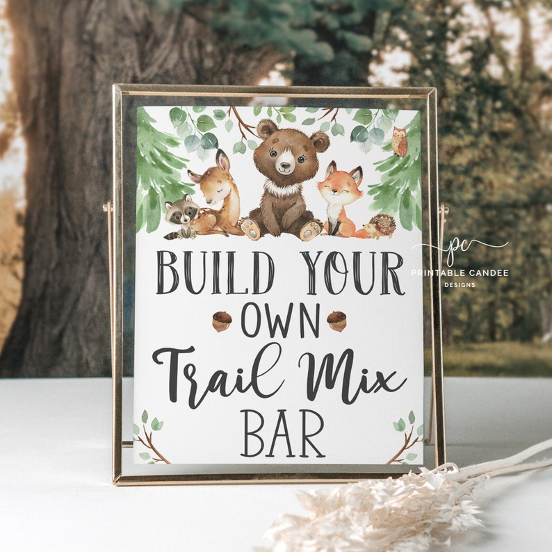May include: A printable sign with a woodland theme featuring a bear, fox, deer, raccoon, and hedgehog. The sign says "Build Your Own Trail Mix Bar".
