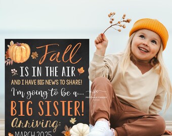 Op de afbeelding: Een krijtbord met een herfstthema dat de komst van een baby aankondigt. Het bord staat er: "De herfst hangt in de lucht en ik heb groot nieuws te delen! Ik ga een... GROTE ZUSTER worden! Aankomst: maart 2025". Een jong meisje in een bruine trui en een oranje muts zit voor het bord en glimlacht.