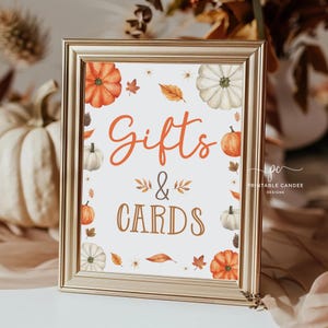 May include: A gold-framed sign with a white background and fall-themed decorations. The sign reads "Gifts & Cards" in brown text with a decorative flourish.