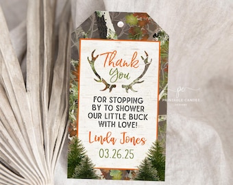 May include: A brown and green camouflage thank you tag with a white background. The tag features a stylised orange deer antler design and the text "Thank You" in orange. The text "For stopping by to shower our little buck with love!" is in orange. The text "Linda Jones 03.26.25" is in orange. The tag is ideal for a baby shower.