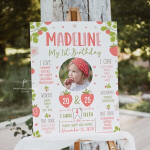 May include: A personalized first birthday sign featuring the name "Madeline" in pink and red. The sign includes a photo, weight of 20 pounds, height of 25 inches, and other details like favorite foods and nicknames. The sign is decorated with strawberries.