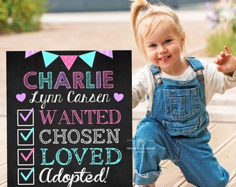 May include: A black chalkboard sign with the text "CHARLIE Lynn Carsen" in pink and blue lettering. The sign also includes a checklist with the words "WANTED", "CHOSEN", "LOVED", and "Adopted!" in pink and blue. The date "JUNE 15, 2025" is written in purple at the bottom of the sign.