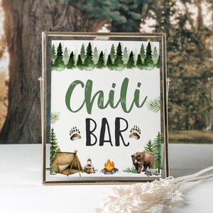 Op de afbeelding: Een rustiek houten bord met een groene aquarel achtergrond van bomen en de tekst "Chili Bar" in groene en zwarte letters. Het bord heeft ook een bruine beer, een kampvuur en een tent.
