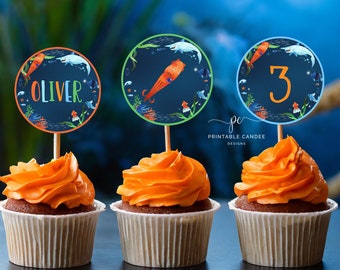 Puede incluir: Tres cupcakes con glaseado naranja cubiertos con toppers redondos de papel azul y verde. Los toppers representan un pulpo, un pez, el nombre "Oliver" y el número "3".