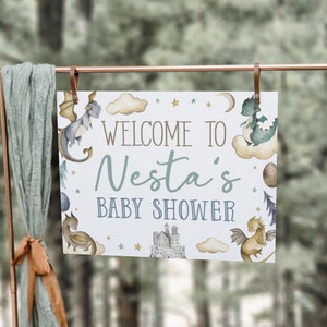 May include: A white baby shower sign with a blue and green dragon illustration, clouds, stars, and the text "Welcome to Nesta's Baby Shower".