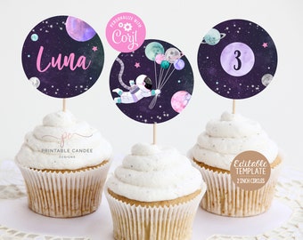 May include: Three cupcake toppers with a space theme. The toppers feature a purple and pink astronaut holding balloons, a purple and pink planet with the number 3, and a purple and pink planet with the name Luna.