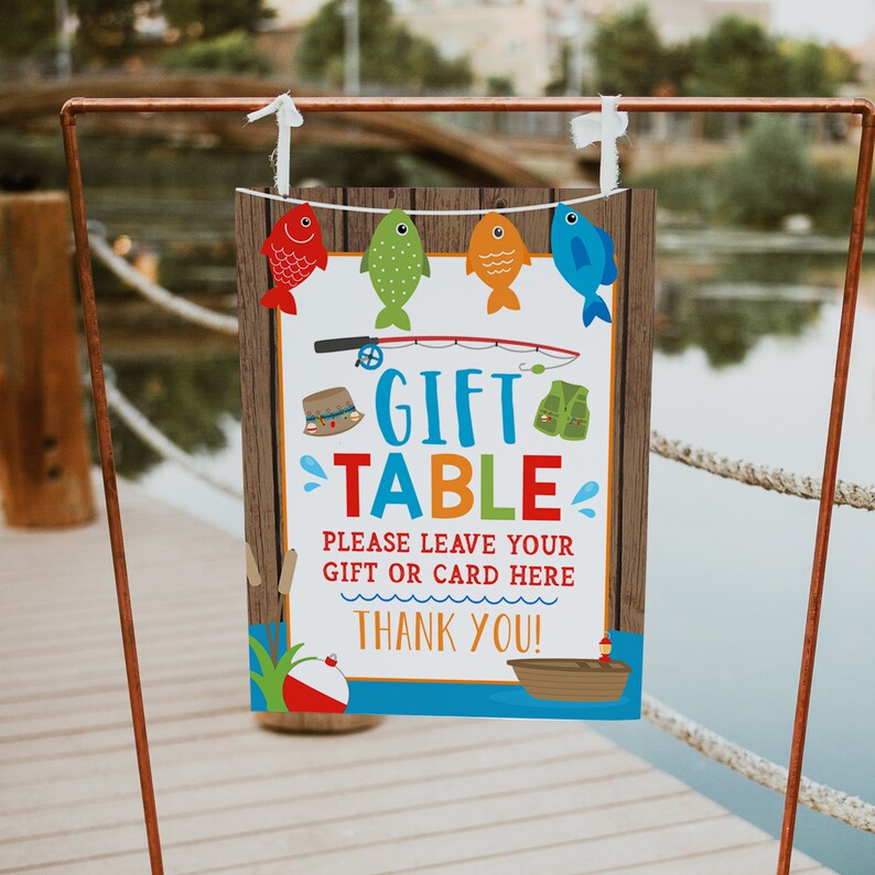 K&ouml;nnte beinhalten: Ein farbenfrohes Schild mit einem Holzhintergrund und dem Text "Gift Table" in blauen und gr&uuml;nen Buchstaben. Das Schild enth&auml;lt auch eine Angelrute, eine Schwimmweste, einen Angelk&ouml;der und ein Boot. Der Text "Please leave your gift or card here" ist in schwarzen Buchstaben geschrieben. Das Schild sagt auch "Thank you!" in schwarzen Buchstaben.