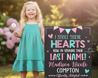 May include: A young girl in a teal dress holds a dandelion, smiling next to a sign. The sign reads "I STOLE THEIR HEARTS, NOW I'M STEALING THEIR LAST NAME!" with the name "Madison Nicole Compton" and the date "August 15, 2026."