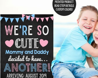 May include: A chalkboard sign with the text "We're so cute Mommy and Daddy decided to have... ANOTHER! Arriving: August 2019". The sign is decorated with pink and blue hearts and bunting. Two children are smiling in the background.