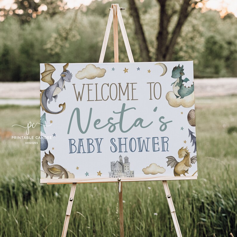 May include: A white welcome sign with blue and gold accents featuring a dragon and castle illustration. The sign reads "Welcome to Nesta's Baby Shower".