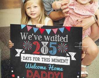 May include: A black chalkboard sign with red, white, and blue decorations. The sign reads "We've waited 285 days for this moment Welcome Home Daddy!"