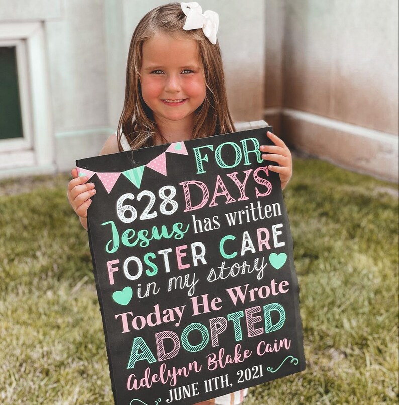 May include: A black chalkboard sign with pink and green decorations announcing that a child has been adopted after 628 days in foster care. The sign reads "FOR 628 DAYS Jesus has written FOSTER CARE in my story Today He Wrote ADOPTED Adelynn Blake Cain JUNE 11TH, 2021".