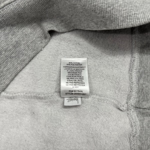 May include: Close-up of a gray sweatshirt interior with a white tag. The tag includes washing instructions in French and the composition of the fabric: 80% cotton and 20% polyester. The tag also includes the style number.