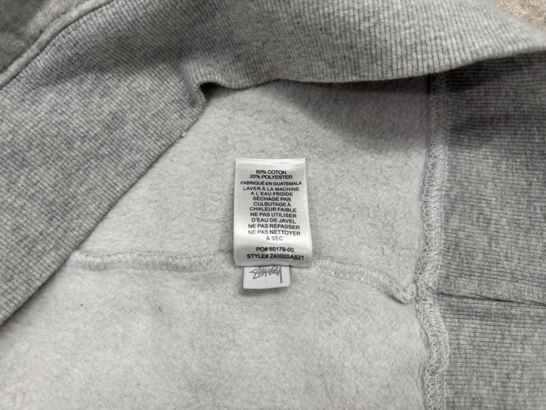 May include: Close-up of a gray sweatshirt interior with a white tag. The tag includes washing instructions in French and the composition of the fabric: 80% cotton and 20% polyester. The tag also includes the style number.