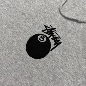 May include: A gray hoodie featuring a black graphic of an eight ball and the Stussy logo. The hoodie has a drawstring hood and a kangaroo pocket. The design is printed on the front of the hoodie.