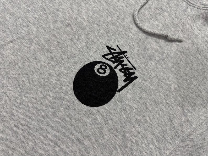 May include: A gray hoodie featuring a black graphic of an eight ball and the Stussy logo. The hoodie has a drawstring hood and a kangaroo pocket. The design is printed on the front of the hoodie.